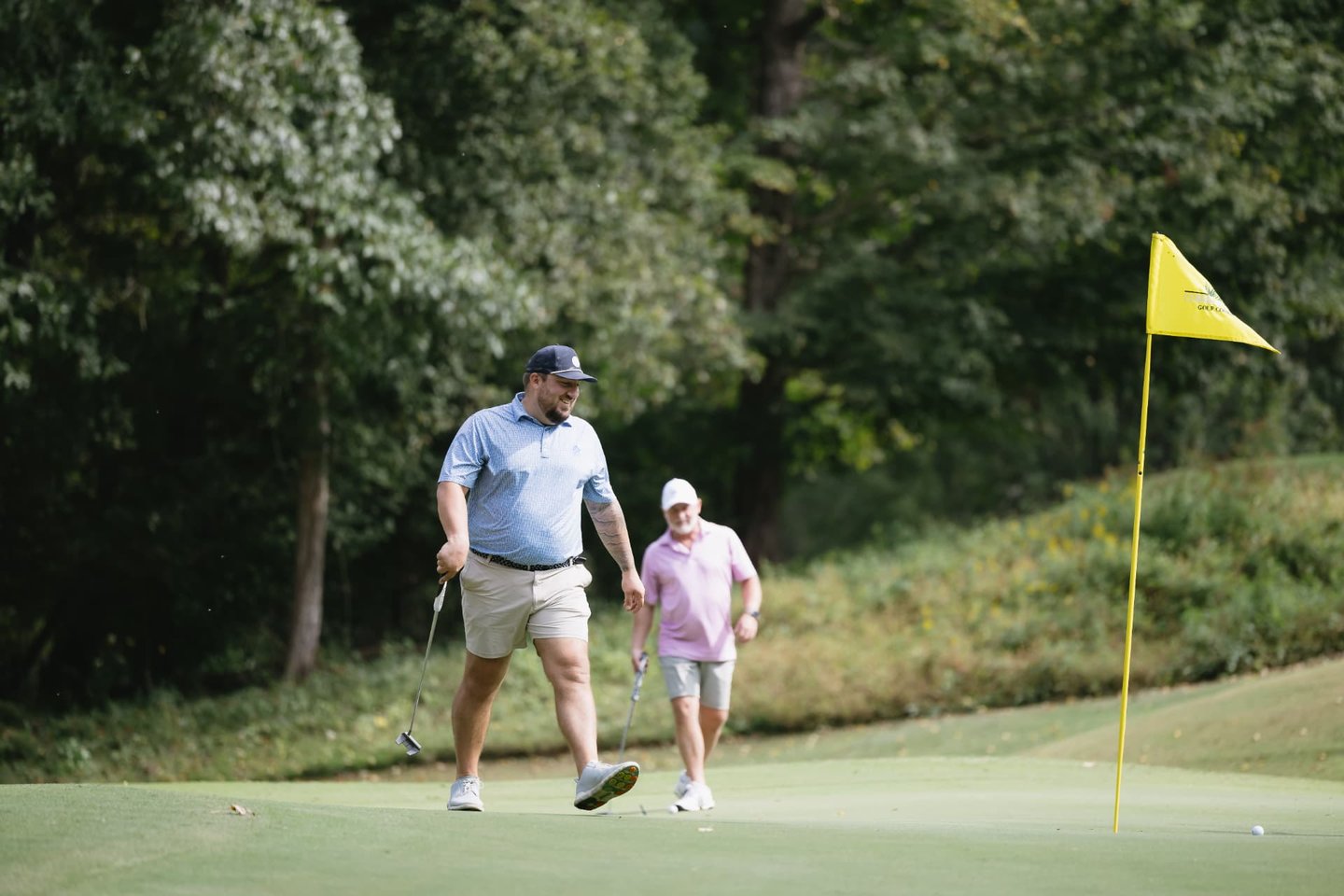 A golfer reacting with a laugh after a putt slides past the hole, a playing partner walking up behind him.