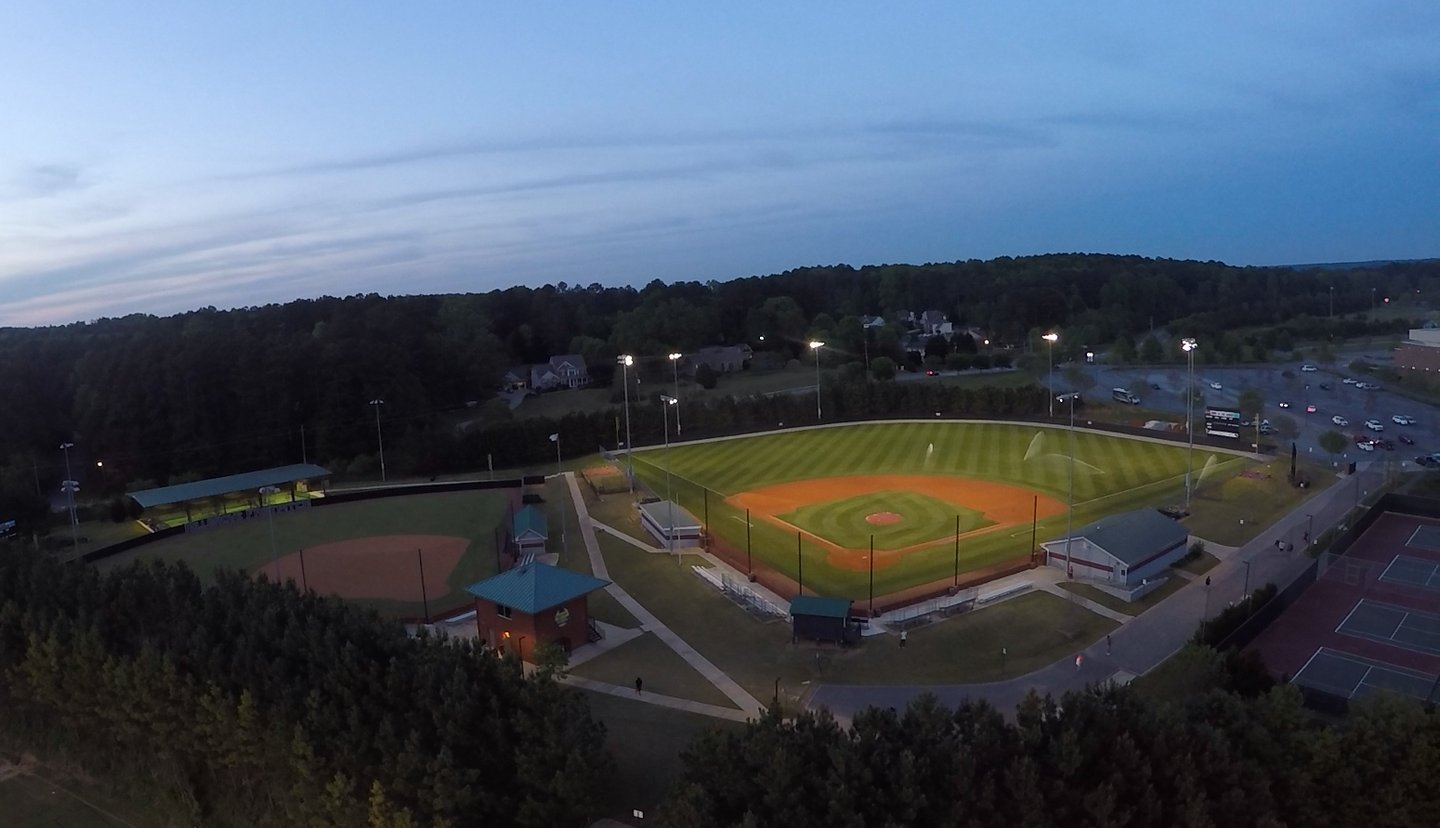 Aerial view at dusk of the Hillgrove baseball and softball complex with the lights on and sprinklers running on the infield.